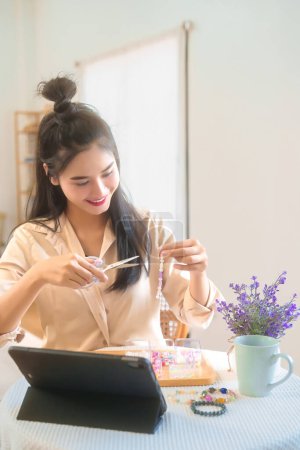 Téléchargez les photos : Jeunes femmes asiatiques regarder bricolage accessoires tutoriel sur tablette numérique tout en utilisant des ciseaux et ficelle perles colorées sur le fil à faire bracelet à la main et faire style de vie passe-temps à la maison atelier. - en image libre de droit