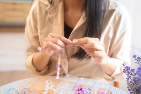 Téléchargez les photos : Les jeunes femmes asiatiques cueillant des perles dans la boîte et ficelant des perles colorées sur le fil à faire bracelet à la main tout en regardant tutoriel accessoires bricolage sur tablette et faire style de vie passe-temps à la maison. - en image libre de droit