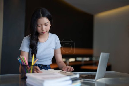 Téléchargez les photos : Asiatique adolescent étudiant femme lire un livre et écrire des notes pour faire des devoirs tout en regardant la vidéo de conférence leçon en ligne sur ordinateur portable numérique pour étudier et apprendre sur la connaissance de l'éducation dans le café. - en image libre de droit