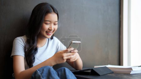 Téléchargez les photos : Asiatique adolescent femme étudiante en utilisant smartphone pour surfer sur les médias sociaux et discuter avec des amis avec bonheur après avoir étudié et lire la paperasse pour en apprendre davantage sur la connaissance de l'éducation dans le café. - en image libre de droit