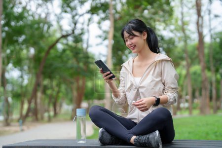 Téléchargez les photos : Jeunes femmes asiatiques sportives portant un écouteur pour écouter de la musique et vérifier l'application de fitness sur smartphone tout en se préparant à courir et à s'entraîner pour un mode de vie sain le matin au parc extérieur. - en image libre de droit