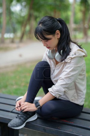 Téléchargez les photos : Jeunes femmes asiatiques sportives assises sur un banc et attachant des lacets en cours d'exécution pour se préparer à la formation de jogging pendant l'entraînement et l'exercice pour un mode de vie sain de bien-être le matin au parc extérieur. - en image libre de droit