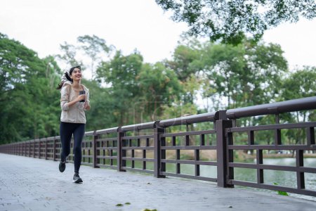 Téléchargez les photos : Jeunes coureuses asiatiques portant des écouteurs et utilisant un smartphone pour écouter la musique avec bonheur tout en faisant du jogging en plein air pour s'entraîner et faire de l'exercice pour un mode de vie sain dans le parc naturel. - en image libre de droit