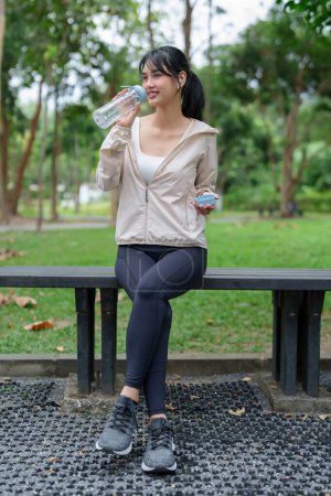 Téléchargez les photos : Les jeunes femmes asiatiques sportives assis pour se reposer sur le banc et boire de l'eau douce avec le bien-être de la santé après avoir couru la formation et l'entraînement d'exercice pour un mode de vie sain le matin au parc extérieur. - en image libre de droit