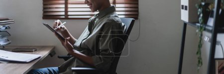 Téléchargez les photos : Femme d'affaires afro-américaine lisant des données financières et prenant des notes sur tablette sur le lieu de travail. - en image libre de droit