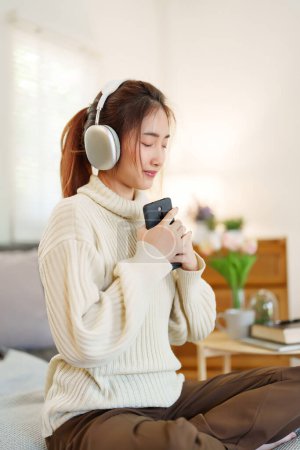 Téléchargez les photos : Jeunes femmes asiatiques en chandail portant un casque pour écouter de la musique et respirer tout en étant assis sur un canapé confortable pour se détendre et faire de l'activité pour passer du temps avec un style de vie lent à la maison. - en image libre de droit