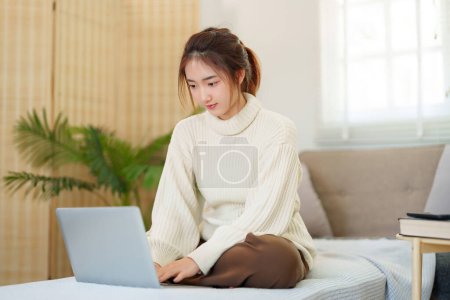 Téléchargez les photos : Jeunes femmes asiatiques en pull assis sur le canapé pour profiter avec surf divertissement sur les médias sociaux sur ordinateur portable tout en se relaxant et en faisant de l'activité pour passer du temps avec style de vie lent à la maison. - en image libre de droit