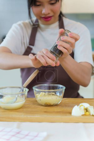 Téléchargez les photos : Femmes asiatiques ajoutant de la mayonnaise et du poivre dans des œufs durs hachés pour mélanger la vinaigrette de salade tout en préparant les ingrédients pour cuisiner le petit déjeuner et faire un mode de vie alimentaire sain dans la cuisine à la maison. - en image libre de droit