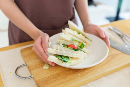 Téléchargez les photos : Gros plan mains de femmes asiatiques tenant une assiette de délicieux sandwich après avoir fait un sandwich avec salade d'œufs durs et légumes tout en cuisinant le repas du petit déjeuner et en faisant un mode de vie alimentaire sain dans la cuisine. - en image libre de droit