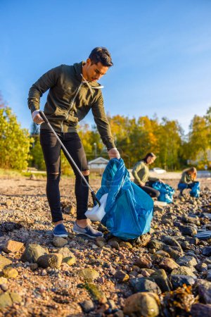 Photo for Full length portrait of young man picking up garbage in bag. Multi-ethnic volunteers are cleaning beach against sky. They are protecting environmental from damage. - Royalty Free Image