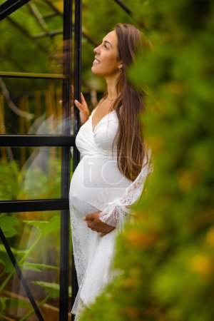 A serene pregnant woman in a white dress standing on a balcony, surrounded by lush greenery, showcasing the beauty and calm of pregnancy.