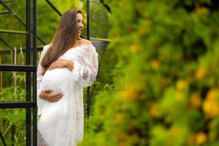 A serene pregnant woman in a white dress cradles her belly while standing in a vibrant, green garden, exuding happiness and anticipation.