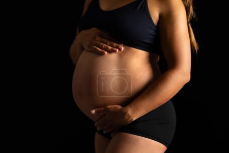 Téléchargez les photos : Gros plan d'une femme enceinte en tenue de sport, tenant sa bosse de bébé sur un fond sombre, dépeignant la maternité et l'anticipation. - en image libre de droit