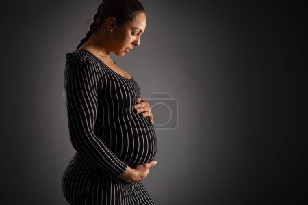Téléchargez les photos : Une femme enceinte sereine en robe rayée, embrassant doucement son ventre sur un fond sombre. Capte la beauté et l'anticipation de la maternité. - en image libre de droit