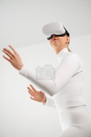 A young woman dressed in white interacts with a virtual environment using a VR headset. The minimalistic background highlights the futuristic concept and technological innovation.