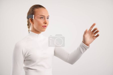Une jeune femme interagit avec la technologie futuriste portable tout en portant des lunettes de sécurité et une tenue blanche moderne, mettant l'accent sur l'innovation et l'interaction numérique dans un environnement de studio neutre.