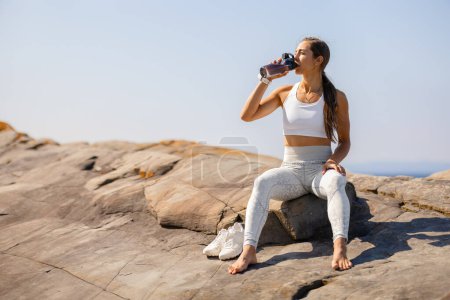 Fit young woman in sportswear hydrates by drinking water while sitting on a rocky coastal landscape. Capturing a moment of well-deserved rest under a clear sky.