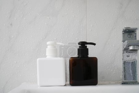 Zwei quadratische Flaschen, eine weiße und eine braune, mit schwarzen Pumps, sitzen auf einem nassen Waschbecken vor einer weiß gekachelten Wand.