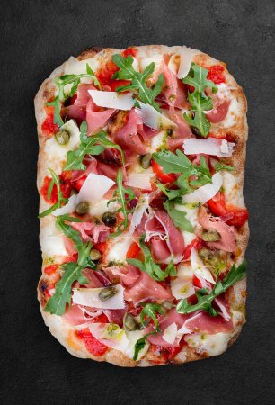 Sorrentina-Pizza mit Schinken, Rucola, Kapern, Pelati-Sauce, Pesto. Römische Pizza rechteckig auf dunklem Hintergrund