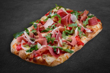 Sorrentina pizza with prosciutto, arugula, capers, pelati sauce, pesto. Roman pizza rectangular on dark background