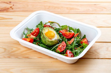 Baked egg in avocado with lettuce and tomatoes. Healthy diet. Takeaway food.  On a wooden background.