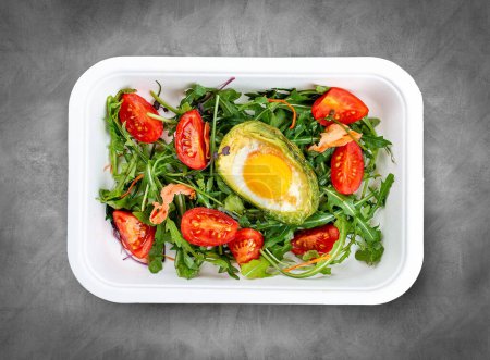 Baked egg in avocado with lettuce and tomatoes. Healthy diet. Takeaway food.  Top view, on a gray background.