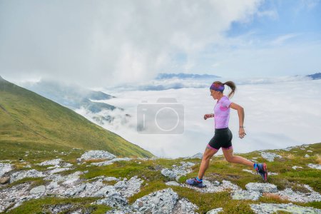 Téléchargez les photos : Femme montant une montagne avec des rochers et de l'herbe vert profond avec une mer de nuages blancs en arrière-plan. - en image libre de droit
