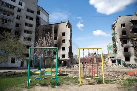 Foto de BORODYANKA, UCRANIA - 27 de abril de 2022: Guerra de Rusia contra Ucrania. Niños balancean cerca de edificios residenciales destruidos después del bombardeo de la ciudad por aviones militares rusos, 27 de abril, 2022. - Imagen libre de derechos
