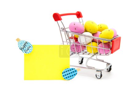 Grußkarte und Einkaufswagen mit bunten glitzernden Ostereiern auf weißem Hintergrund. Frohe Ostern! Freiraum für Text. Kauf von Produkten für den Urlaub.