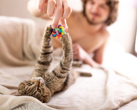 Young man playing with his small cat in bed
