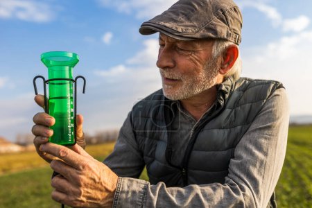 Téléchargez les photos : Un agriculteur âgé regarde un pluviomètre pour mesurer la pluie dans un champ ou un jardin - en image libre de droit