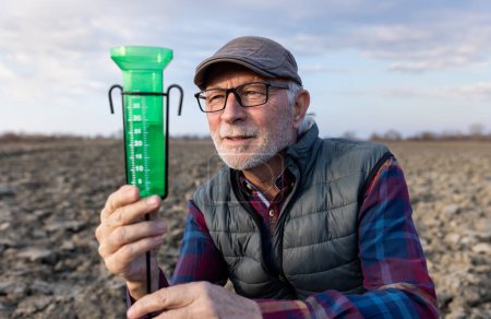 Téléchargez les photos : Un agriculteur âgé examine un pluviomètre pour mesurer la pluie dans un champ - en image libre de droit