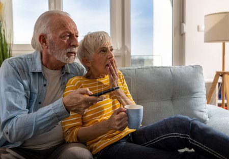 Téléchargez les photos : Couple de personnes âgées regardant des scènes effrayantes dans un film sur le canapé à la maison. Homme mûr tenant télécommande - en image libre de droit