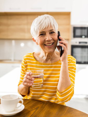 Photo for Pretty mature woman talking on mobile phone and holding glass of water at dining table at home - Royalty Free Image