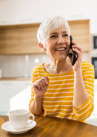 Photo for Cheerful mature woman talking on mobile phone and drinking coffee at home at dining table - Royalty Free Image