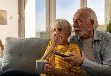 Téléchargez les photos : Couple de personnes âgées regardant des scènes effrayantes dans un film sur le canapé à la maison. Homme mûr tenant télécommande - en image libre de droit