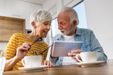 Seniorenpaar sitzt am Esstisch und schaut auf Tablet, surft im Internet 