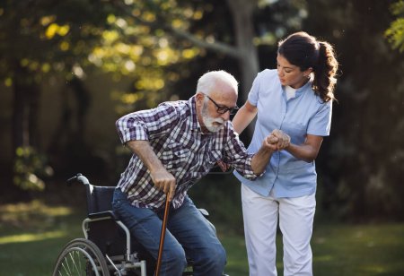 Téléchargez les photos : Jeune femme aidante aidant l'homme âgé à se lever du fauteuil roulant dans le parc - en image libre de droit
