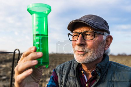 Téléchargez les photos : Senior farmer looking at water gauge for rain measuring in field - en image libre de droit