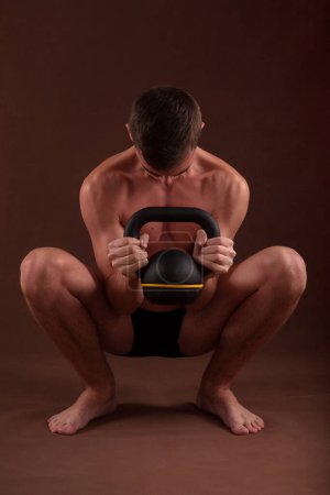 Photo for Handsome muscular adolescent shirtless boy training in deep squat exercise with kettlebell. - Royalty Free Image