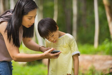 Photo for Beautiful Asian Mom showing to her son something on her phone or to teaching and leaning in nature. - Royalty Free Image
