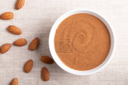 Brown almond butter in a white bowl on linen fabric. Smooth food paste made from grinding almonds into nut butter. Puree of the nuts of Prunus dulcis with nutty flavor, and a good source of protein.
