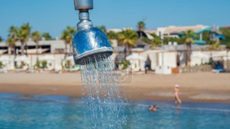 Photo for Outdoor refreshing shower head spraying water to take shower on the pier. Blurry beach and swimming people in the background. - Royalty Free Image