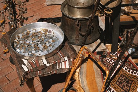 Vintage coins in a metal tray, rusty old cauldron and pots, key chains, rug and other goods in a shop at bazaar. 