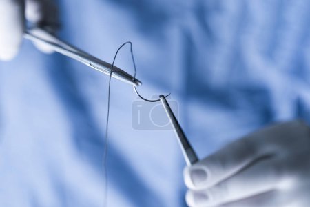 Photo for Close-up of a clinician's hands in blue scrubs meticulously handling surgical sutures. Medical professional in scrubs handling sutures. Operator holding half circle needle with forceps and needle holder during operation. - Royalty Free Image