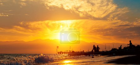 Panoramic summer background with silhouettes of people by the sea.  Beach-goers at the seashore during golden hour in summer in Antalya, Turkey.