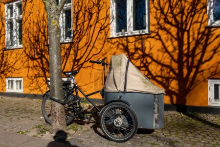 Téléchargez les photos : Copenhague, Danemark Un vélo cargo est verrouillé à un arbre contre une façade orange vif. - en image libre de droit