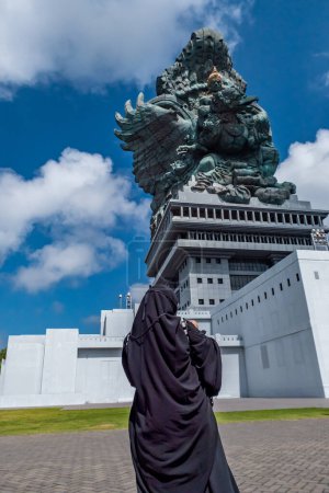 Photo for Bali, Indonesia,  An Arab woman in a veil visits the   GWK Cultural Park outside of Denpasar with statues of Hindu gods, standing under the GWK statue - Royalty Free Image