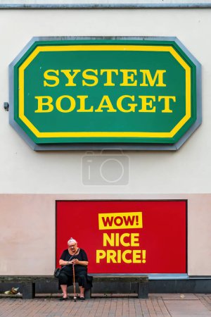 Photo for Stockholm, Sweden June 20 2023 A senior woman sits on a bench on the main square in the Skarholmen suburb under a sign for the System Boloaget, the state run liquor store. - Royalty Free Image