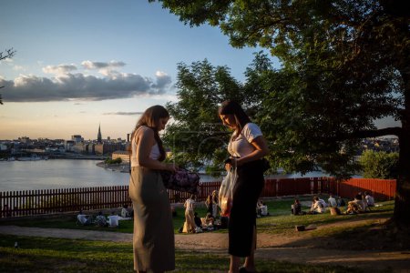 Photo for Stockholm, Sweden June 24, 2023 People sitting in the grass at sunset at Midsummer at the landmark Ivar Lo Park on Sodermalm. and two women packing up their picnic blanket. - Royalty Free Image